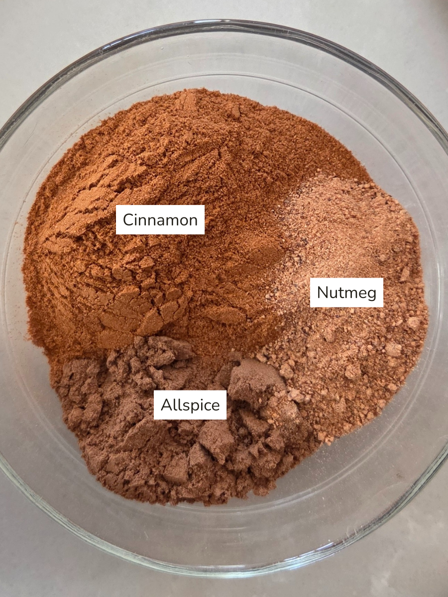 Overhead view of a glass bowl with three ground spices labeled—cinnamon, nutmeg, and allspice—ready to be mixed into homemade apple pie spice blend.