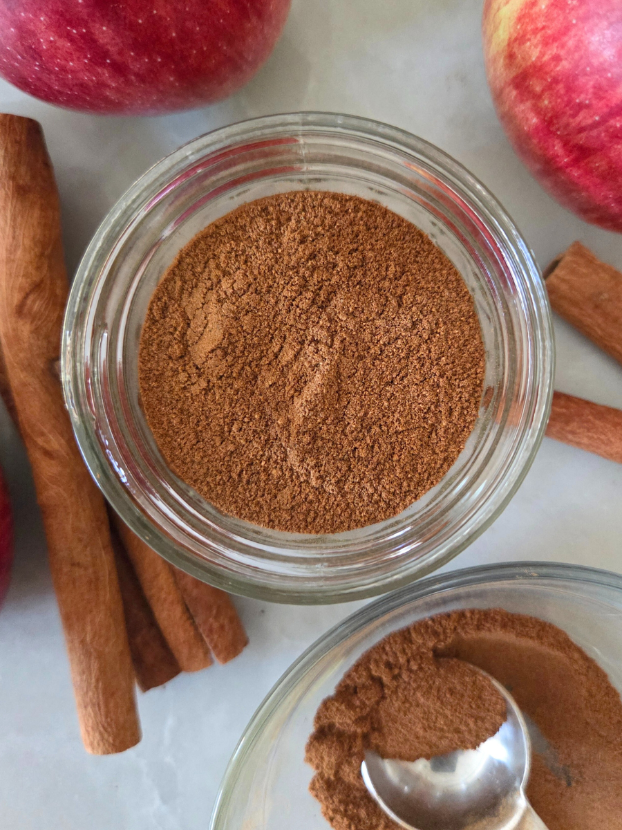 Homemade apple pie spice in a small glass jar surrounded by fresh apples and cinnamon sticks, with a spoonful of spice in a separate bowl.