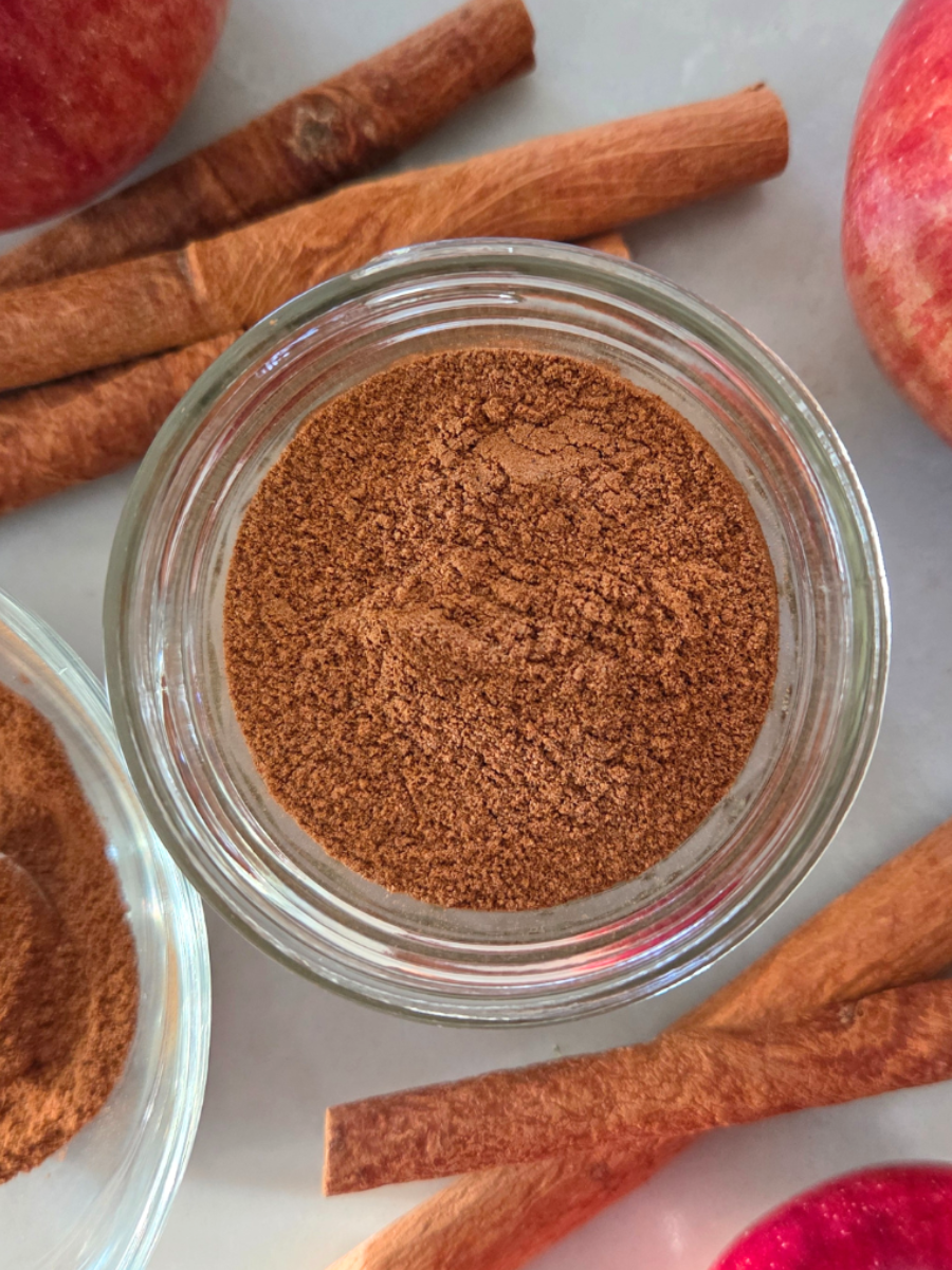 Homemade apple pie spice in a small glass jar with cinnamon sticks and fresh red apples on a neutral background, perfect for fall baking.